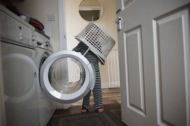 Verspielter Junge, der einen Korb auf dem Kopf trägt, während er zu Hause an der Waschmaschine steht - CAVF15739