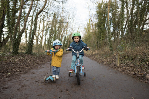 Portrait of siblings riding push scooter and bicycle on road against sky - CAVF15740