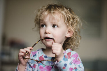 Close-up of girl with eyeglasses in mouth looking away while standing at home - CAVF15743