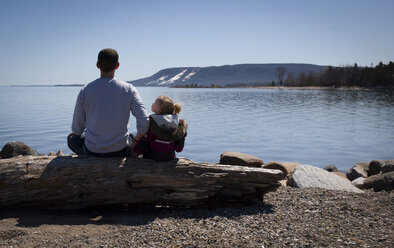 Rear view of father and daughter sitting on log at lakeshore against clear sky during sunny day - CAVF15757