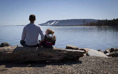 Rückansicht von Vater und Tochter, die auf einem Baumstamm am Seeufer sitzen, gegen einen klaren Himmel an einem sonnigen Tag - CAVF15757