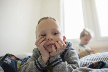 Thoughtful boy looking away while lying on bed with sister in background - CAVF15779