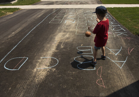 Rückansicht eines Jungen, der mit einem Ball spielt, während er auf der Straße steht und mit Kreide zeichnet - CAVF15802