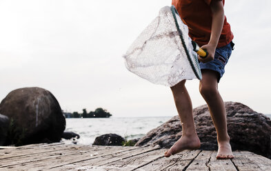 Low section of boy with butterfly fishing net standing on pier against clear sky - CAVF15819