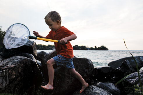 Verspielter Junge läuft auf Felsen, während er ein Schmetterlingsfischernetz gegen das Meer und den klaren Himmel hält - CAVF15821