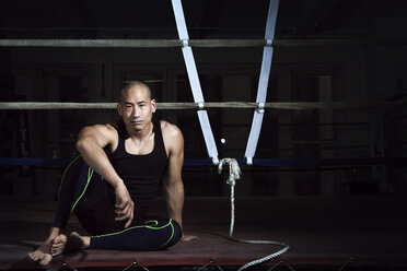 Selbstbewusster Sportler sitzt auf einer Bank vor einem Boxring im Fitnessstudio - CAVF15872