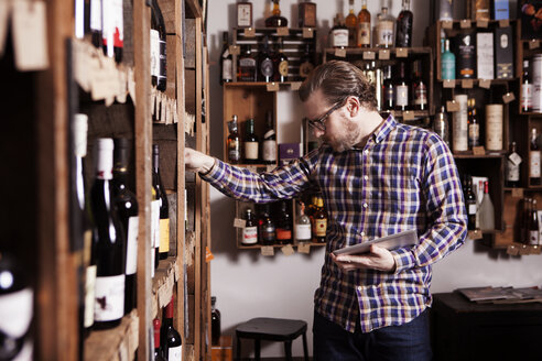 Männlicher Kleinunternehmer, der eine Weinflasche untersucht, während er einen Tablet-Computer im Geschäft hält - CAVF15905