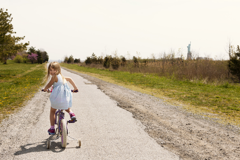 Rückansicht eines Fahrrad fahrenden Mädchens auf einer Straße inmitten eines Feldes, lizenzfreies Stockfoto