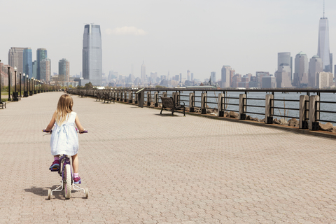 Rückansicht eines Mädchens, das auf der Promenade radelt, mit der Skyline der Stadt im Hintergrund, lizenzfreies Stockfoto
