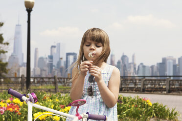 Cute girl blowing dandelion on promenade with city skyline in background - CAVF15973