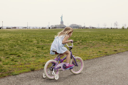 Seitenansicht eines Mädchens, das mit dem Fahrrad auf einer Straße durch ein Feld mit der Freiheitsstatue im Hintergrund fährt - CAVF15976