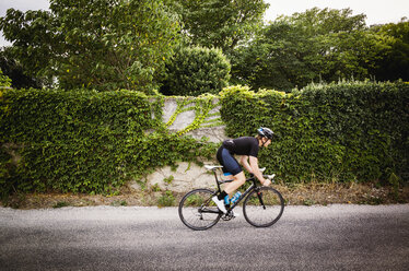 Side view of man cycling on road by ivy covered wall - CAVF16081