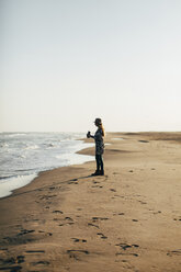 Woman holding camera enjoying at beach against clear sky during sunny day - CAVF16154