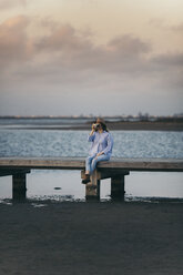 Woman photographing while sitting on pier against cloudy sky during sunset - CAVF16160