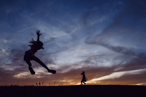 Silhouette Schwestern spielen auf Feld gegen dramatischen Himmel bei Sonnenuntergang, lizenzfreies Stockfoto