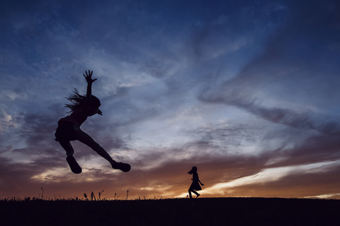 Silhouette sisters playing on field against dramatic sky during sunset stock photo