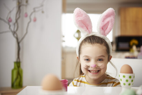 Porträt eines lächelnden Mädchens mit Hasenohren, das am Tisch mit Ostereiern sitzt - ABIF00160