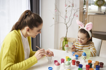 Happy mother and daughter with bunny ears sitting at table painting Easter eggs - ABIF00166