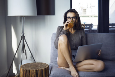 Woman drinking while using laptop computer on sofa - CAVF16353