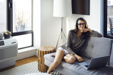 Woman with laptop computer looking away while sitting on sofa - CAVF16355