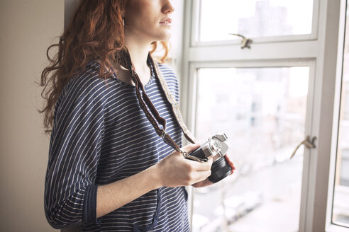 Mittelteil einer Frau mit Kamera, die zu Hause am Fenster steht - CAVF16363