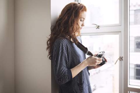 Frau benutzt Digitalkamera, während sie zu Hause am Fenster steht, lizenzfreies Stockfoto