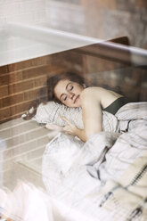 Smiling woman sleeping on bed at home seen through glass - CAVF16407