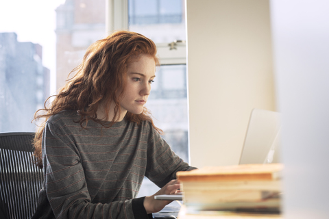 Confident woman using laptop computer while sitting by window at home stock photo