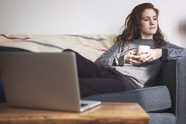 Nachdenkliche Frau mit Kaffeetasse, die wegschaut, während sie sich auf dem Sofa entspannt - CAVF16431