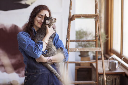 Lächelnder Künstler, der eine Katze umarmt, während er in der Werkstatt steht - CAVF16466