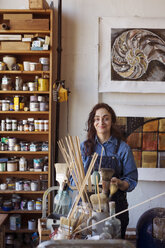 Portrait of smiling artist standing by shelf in workshop - CAVF16470