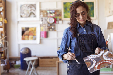 Female artist holding artwork with paintbrush in workshop - CAVF16473