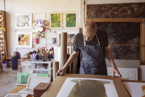 Ältere Künstlerin betrachtet Gemälde am Tisch in der Werkstatt, lizenzfreies Stockfoto