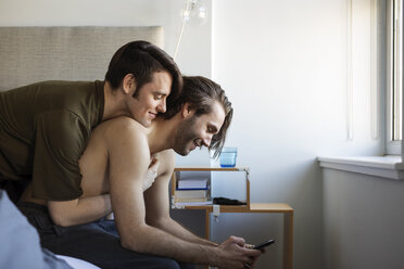 Man embracing boyfriend using mobile phone while sitting on bed - CAVF16507