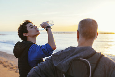 Sohn trinkt Wasser, während er neben seinem Vater am Strand bei Sonnenuntergang steht - CAVF16554
