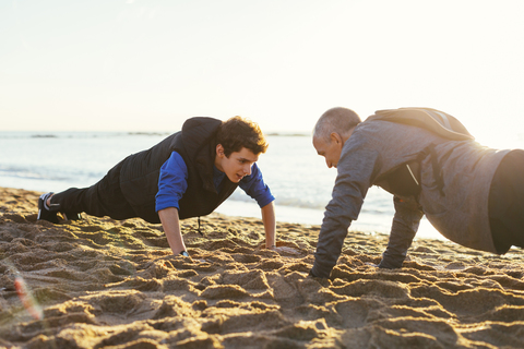 Entschlossener Vater und Sohn machen gemeinsam Liegestütze gegen den klaren Himmel am Strand bei Sonnenuntergang, lizenzfreies Stockfoto