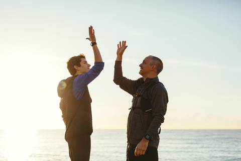 Glücklicher Vater und Sohn mit High-Five gegen Meer und Himmel bei Sonnenuntergang, lizenzfreies Stockfoto