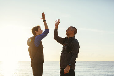 Glücklicher Vater und Sohn mit High-Five gegen Meer und Himmel bei Sonnenuntergang - CAVF16571