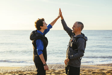 Vater und Sohn geben sich High-Five, während sie bei Sonnenuntergang am Strand vor dem Meer und dem Himmel stehen - CAVF16572