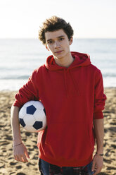Portrait of confident teenage boy holding soccer ball while standing at beach against sea - CAVF16583