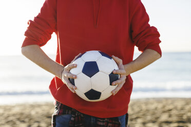 Mittelteil eines Teenagers, der einen Fußball hält, während er am Strand gegen das Meer steht - CAVF16584