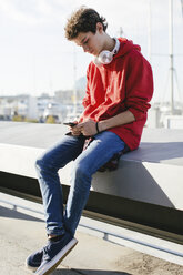 Teenage boy using smart phone while sitting on retaining wall during sunny day - CAVF16605