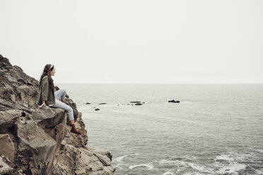 Thoughtful woman sitting on cliff by sea against clear sky - CAVF16657