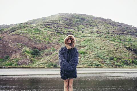 Frau mit Jacke und stehend am nassen Strand vor einem Berg, lizenzfreies Stockfoto