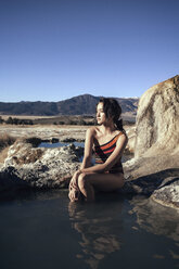 Nachdenkliche Frau sitzt auf einem Felsen bei Bridgeport Hot Springs vor blauem Himmel - CAVF16696