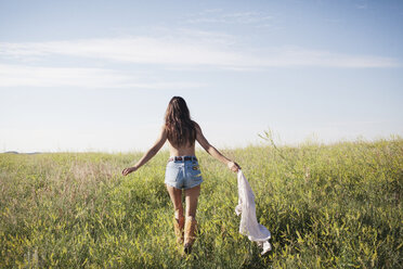 Rückansicht einer Frau, die ein Hemd hält und auf einem grasbewachsenen Feld gegen den Himmel läuft - CAVF16724