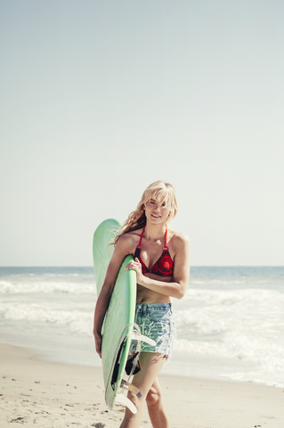 Porträt einer selbstbewussten Frau, die ein Surfbrett trägt, während sie am Strand spazieren geht, lizenzfreies Stockfoto