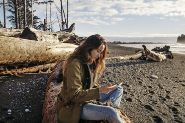 Side view of woman using smart phone while sitting on beach - CAVF16770