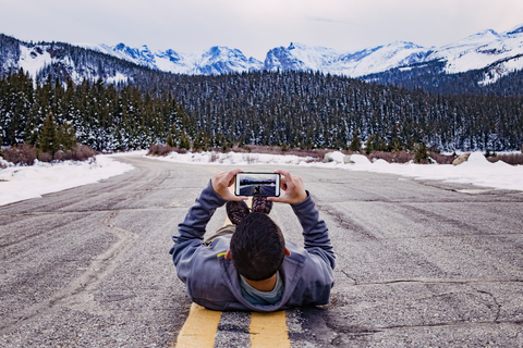 Mann liegt auf der Straße und fotografiert mit Smartphone vor schneebedeckten Bergen, lizenzfreies Stockfoto