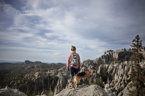 Frau und Hund stehen auf einem Felsen vor Bergen und Himmel, lizenzfreies Stockfoto